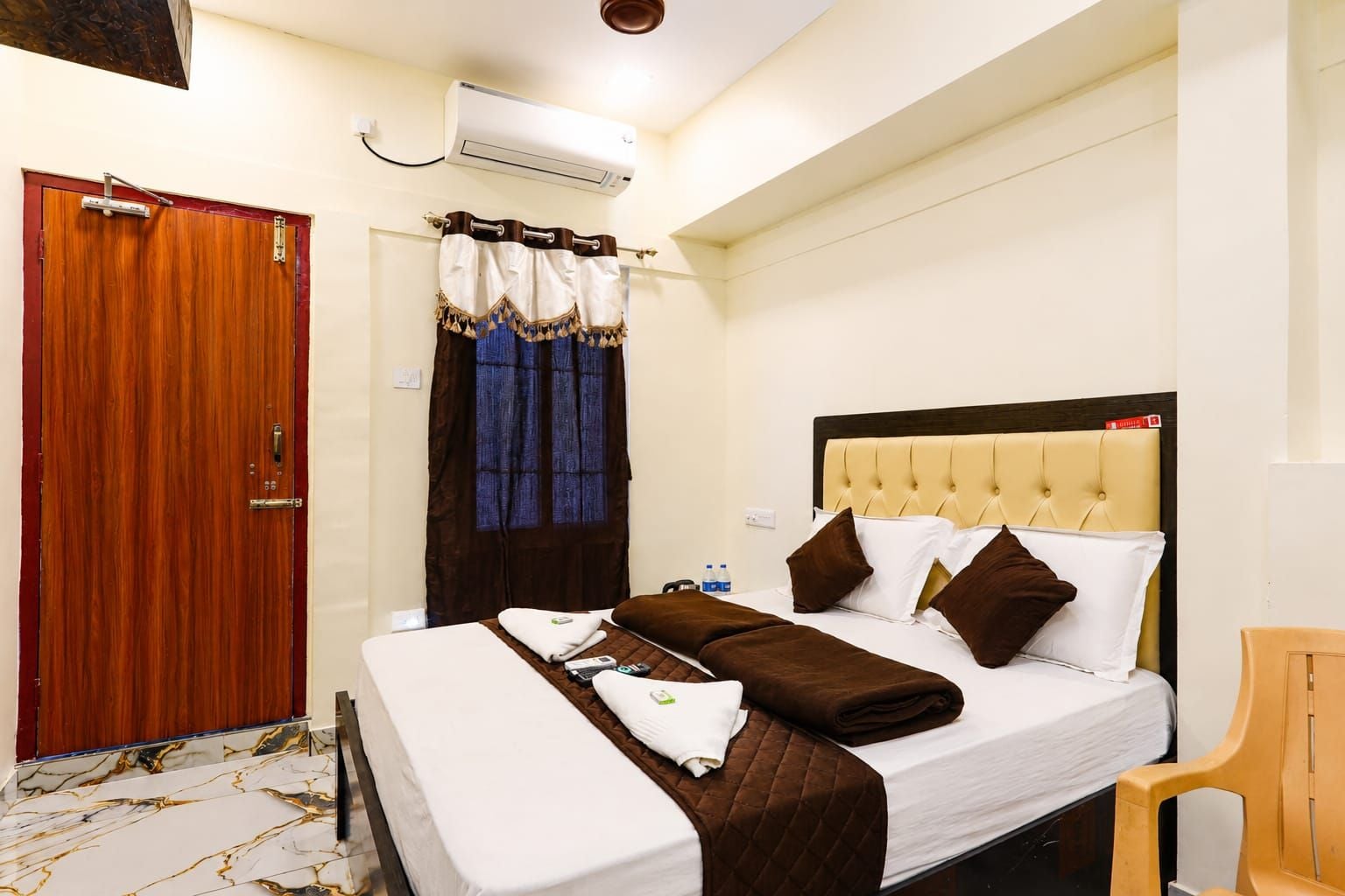 A hotel room with a bed, a wooden door, an air conditioning unit, and a chair.