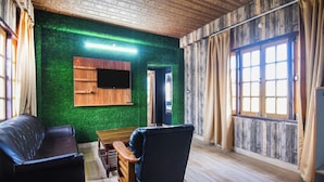 Una habitación con techo de madera, pared verde, mueble de televisión de madera, sofá de cuero negro, banco de madera y un ventanal con cortinas.