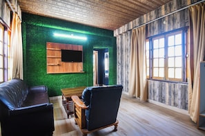 Una habitación con techo de madera, pared verde, mueble de televisión de madera, sofá de cuero negro, banco de madera y un ventanal con cortinas.