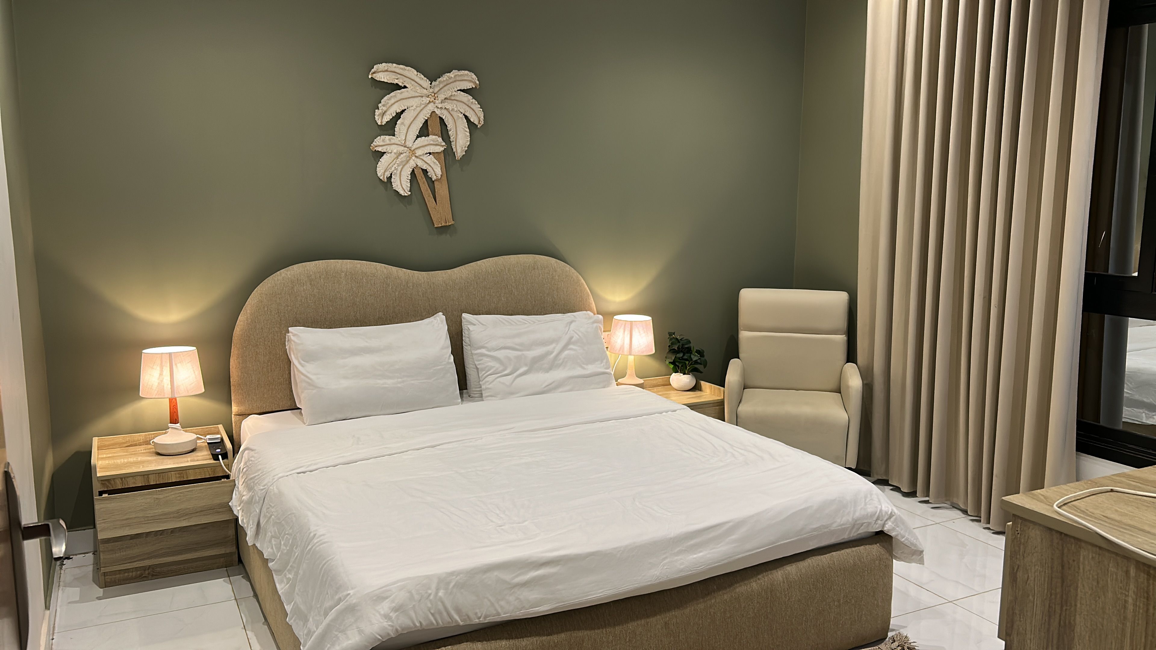 A hotel room with a bed, two bedside tables with lamps, a chair, a plant, and a wall-mounted palm tree decoration.
