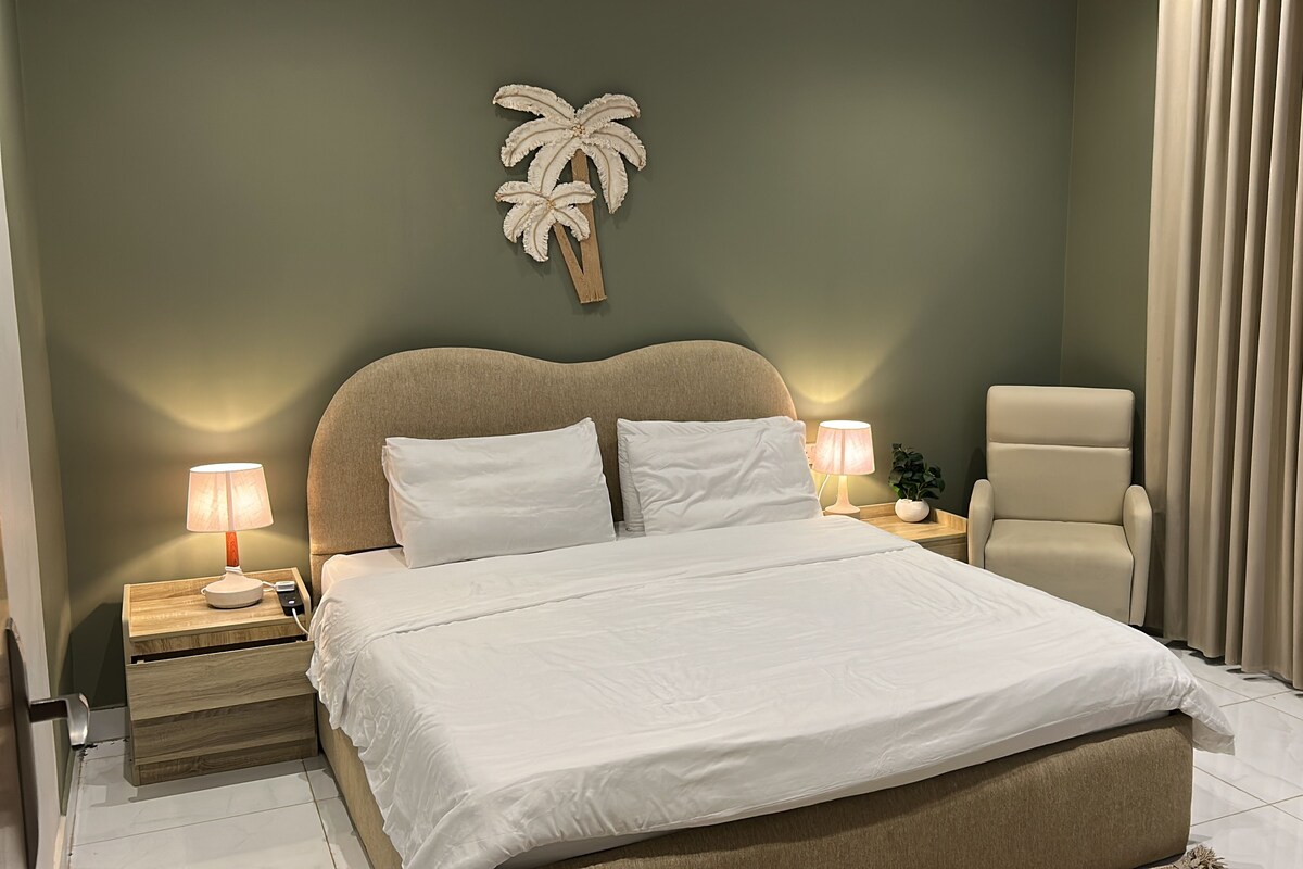 Une chambre d’hôtel comprenant un lit, deux tables de chevet avec des lampes, une chaise, une plante et un décor mural représentant un palmier.