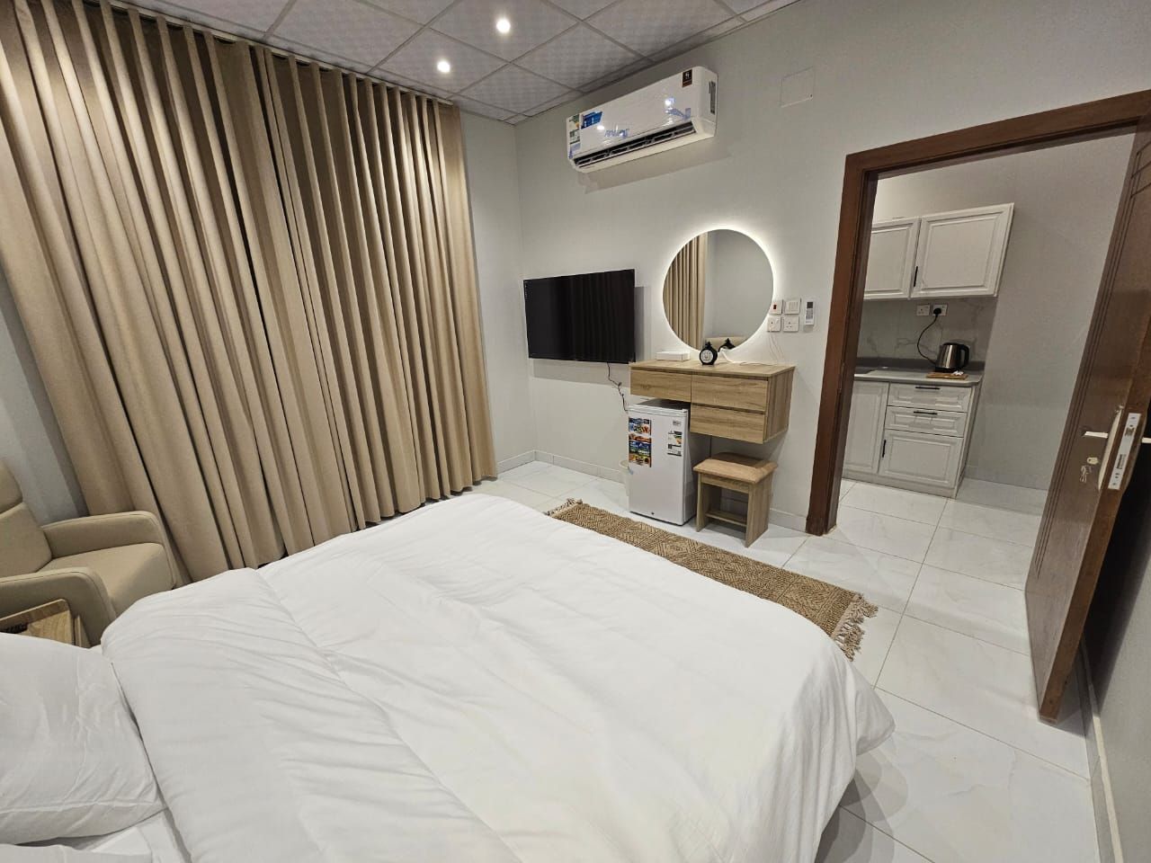 Una habitación de hotel con cama, televisión, minibar, espejo y kitchenette.