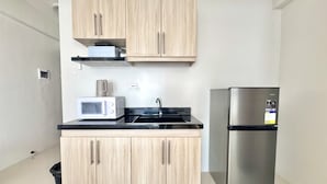 Cozinha compacta com armários de madeira clara, geladeira de aço inoxidável, micro-ondas e bancada preta.