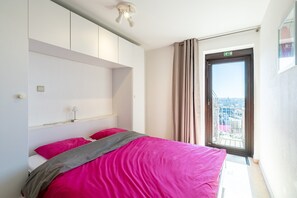 A bedroom with a bed, a bedside table, a lamp, a window with curtains, and a view of the city. - Apartment in Blankenberge Near the Pier (Blankenberge)