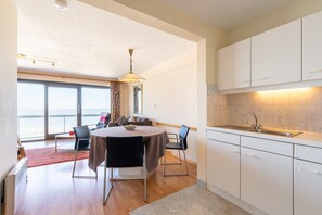 A modern kitchen with white cabinets, a dining area with a round table and chairs, and a large window providing a view of the outdoors. - Apartment in Blankenberge Near the Pier (Blankenberge)