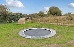 Ein Trampolin in einer grünen, offenen Umgebung mit einem Stein und einer Holzkonstruktion im Hintergrund.