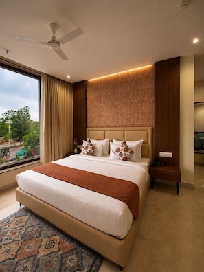 Habitación de hotel con una cama grande, ventilador de techo, mesita de noche y vistas al exterior.