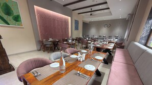 A restaurant interior with multiple dining tables set with white plates, silverware, and a bottle of water. There are cushioned chairs and a sofa in the seating area. - Regenta Place Kota (Kota)