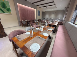 Interior de restaurante con varias mesas dispuestas, con platos blancos, cubiertos y una botella de agua. El área de estar incluye sillas con cojines y un sofá.