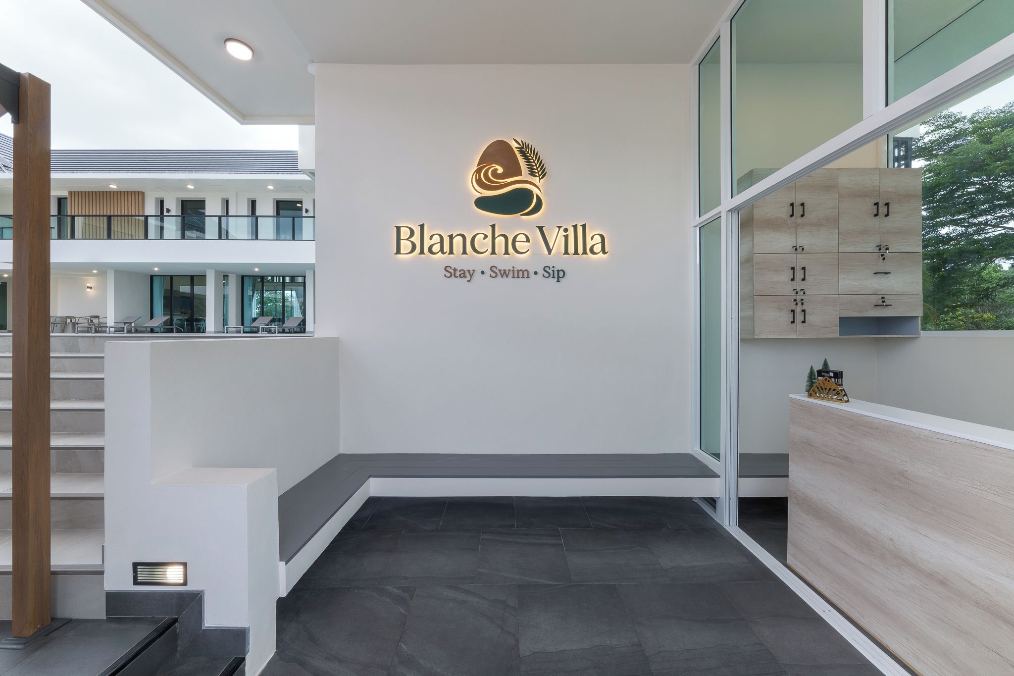 Una moderna zona de recepción de hotel con un logo para Blanche Villa, que muestra el logo y las palabras 'Stay · Swim · Sip'.