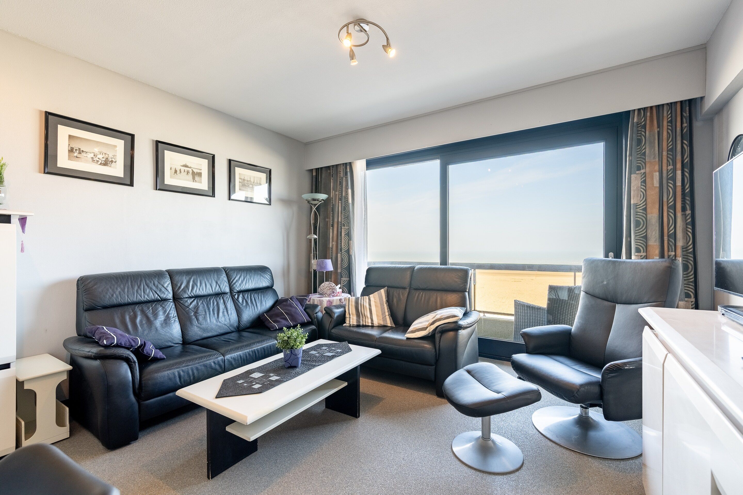 Ein modernes Wohnzimmer mit einer schwarzen Ledersofa, einem passenden Sessel und einem kleinen Couchtisch. An der Wand hängen drei gerahmte Bilder, eine Stehlampe und ein großes Fenster mit Vorhängen.