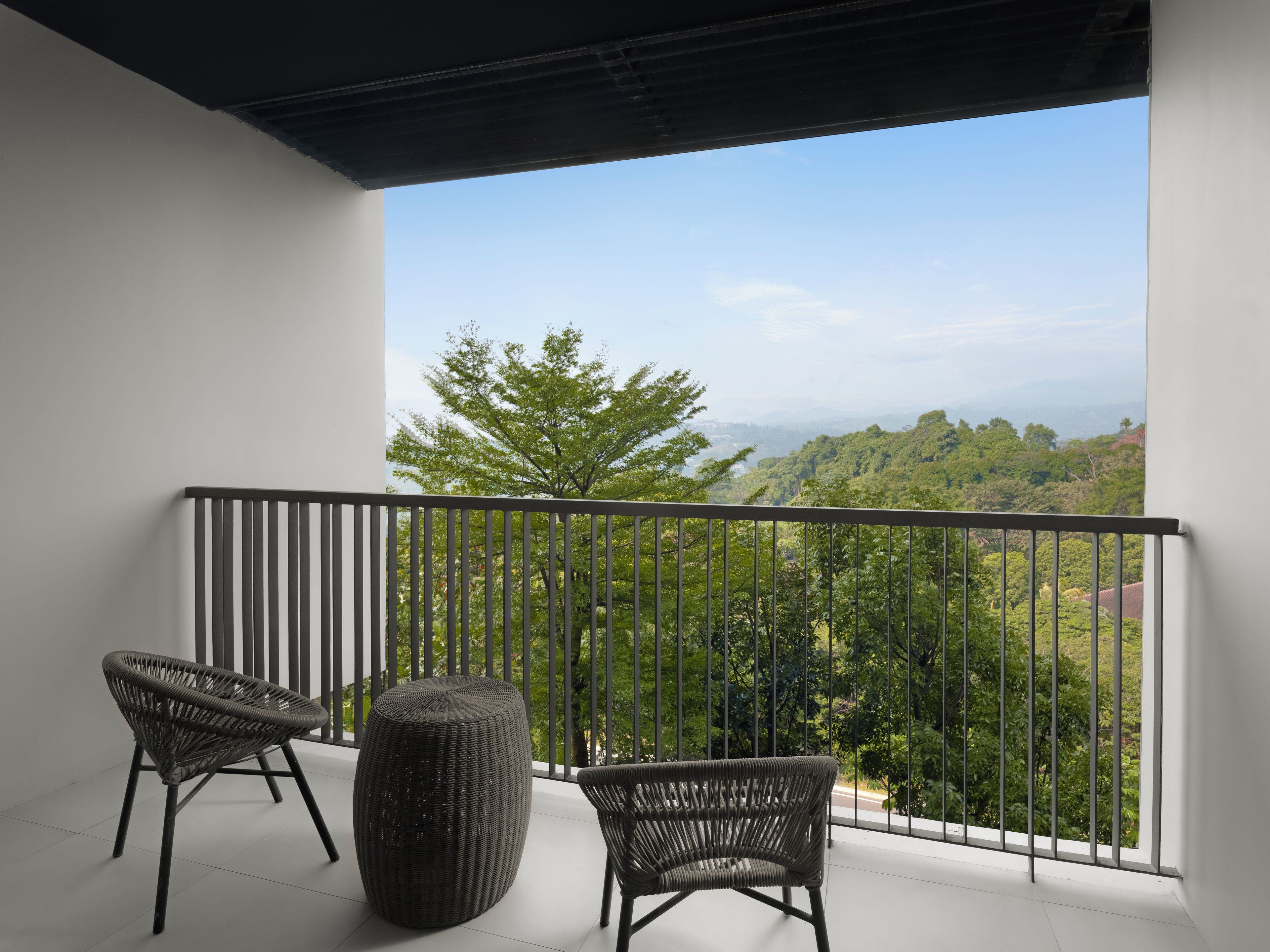A balcony with two wicker chairs and a round ottoman, overlooking a lush green landscape.