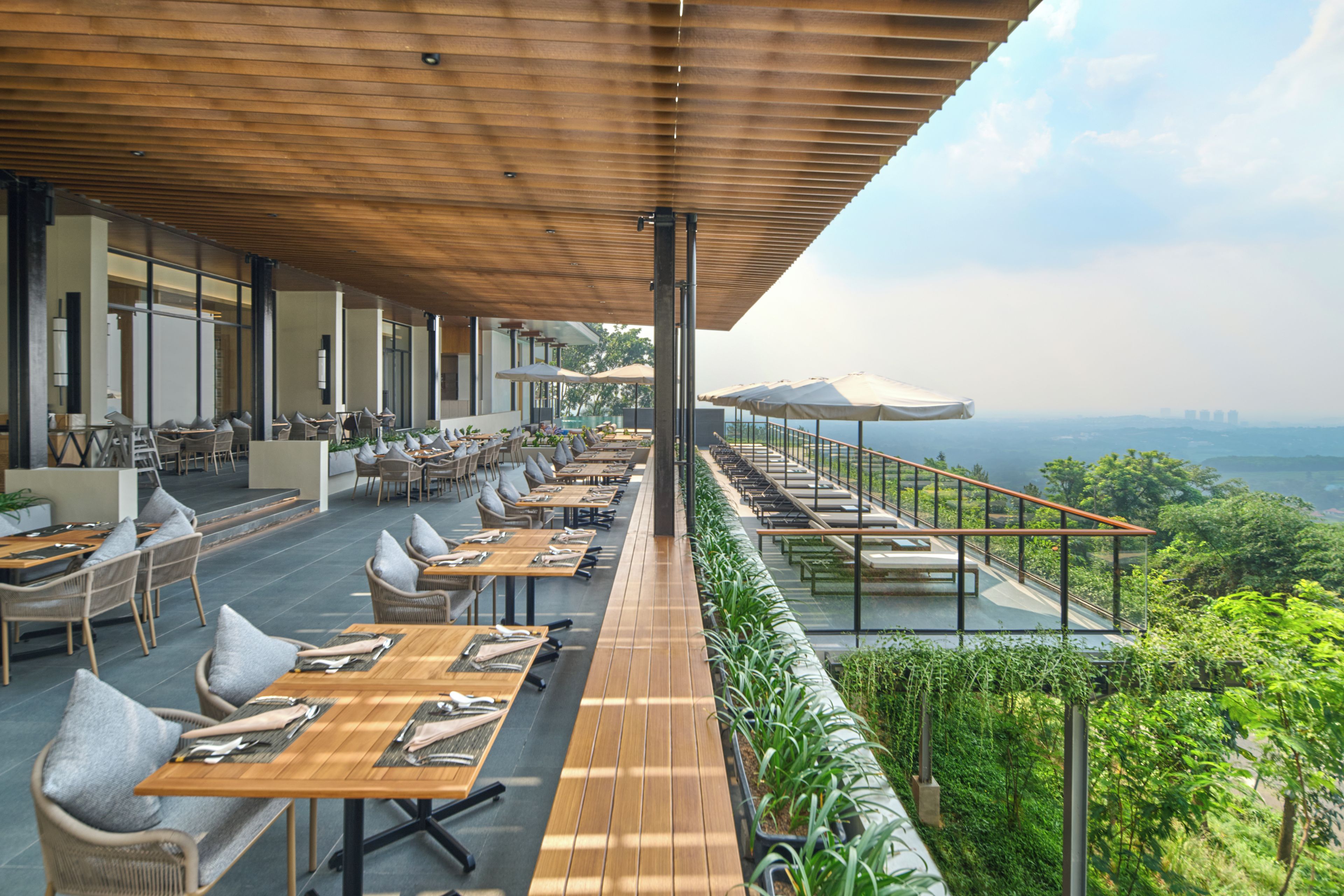 Un área de comedor al aire libre con mesas y sillas de madera, un paseo techado y vistas del paisaje.