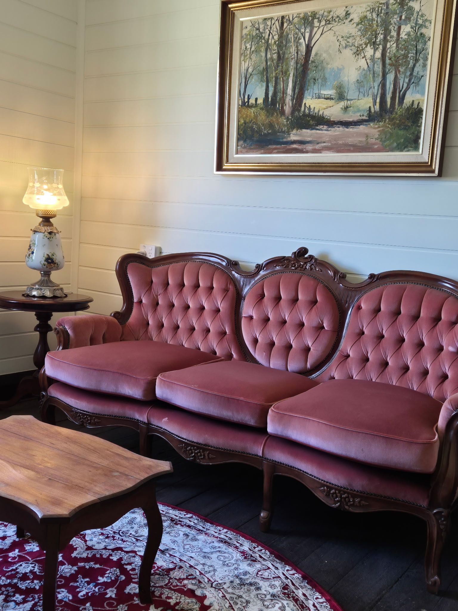A room with a two-seater sofa, a wooden coffee table, a lamp, and a painting on the wall.