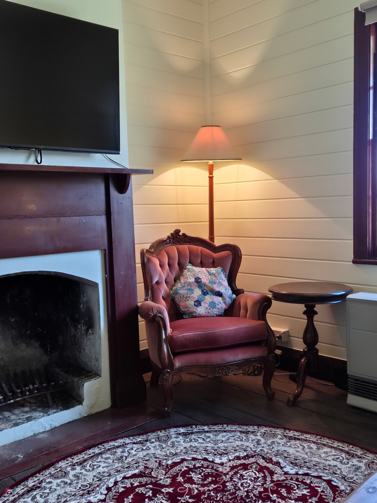 A room with a fireplace, a red armchair with a floral cushion, a small table, and a patterned rug.