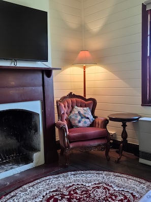 A room with a fireplace, a red armchair with a floral cushion, a small table, and a patterned rug.