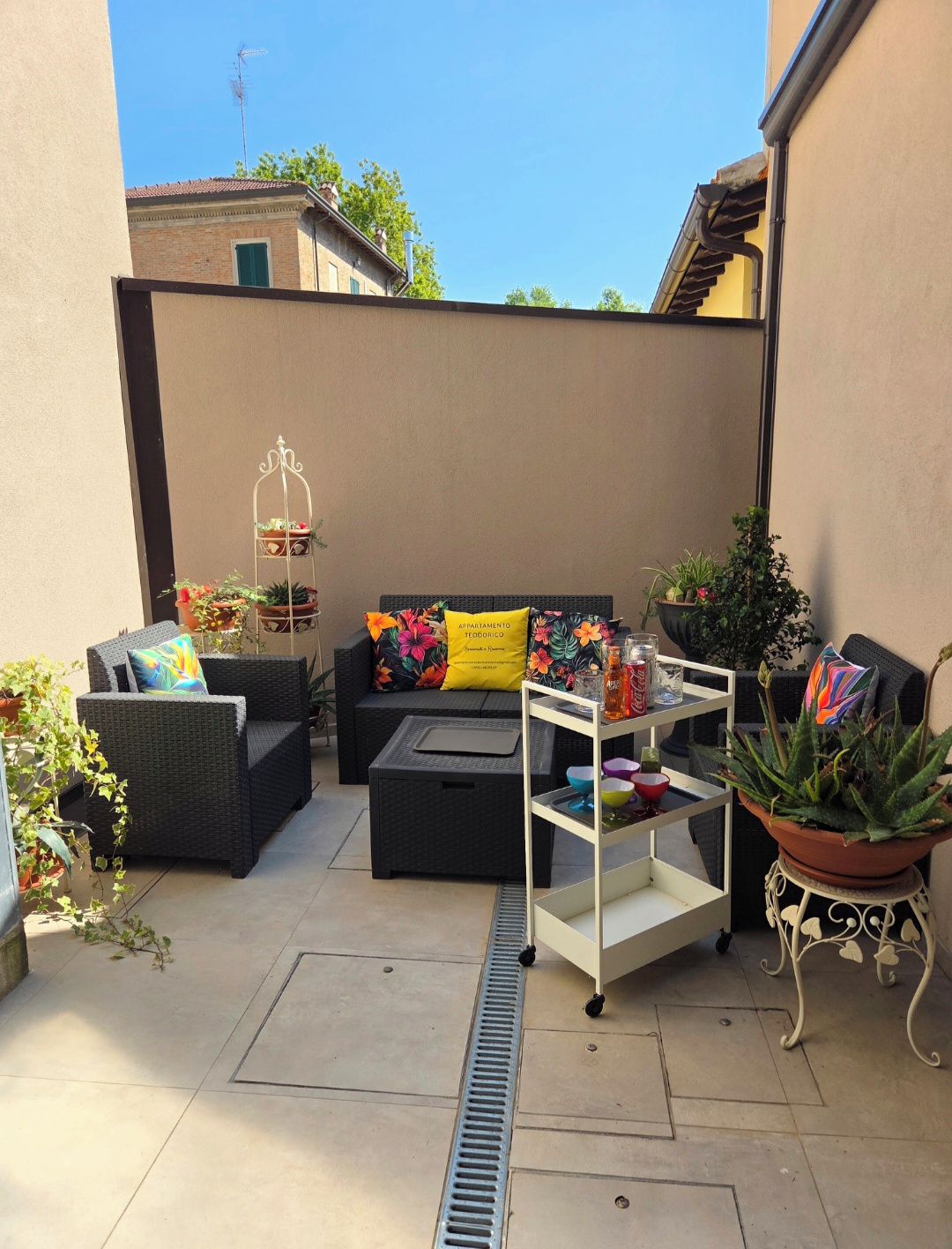 Un patio con mobiliario exterior, un carro metálico blanco y plantas en macetas.