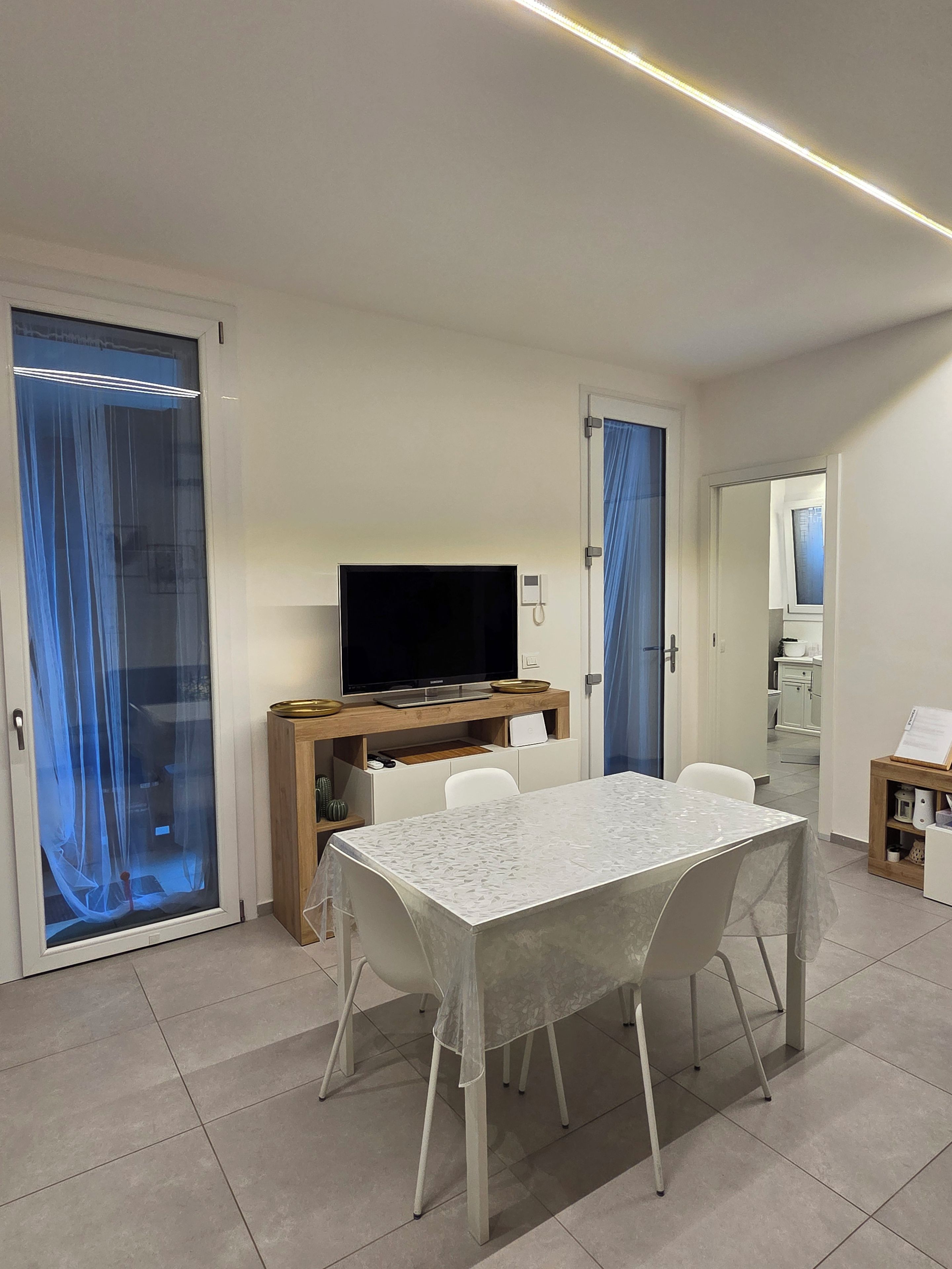 Een moderne woonkamer met een flatscreen televisie op een houten standaard, een eettafel met stoelen, en een keuken in de achtergrond.