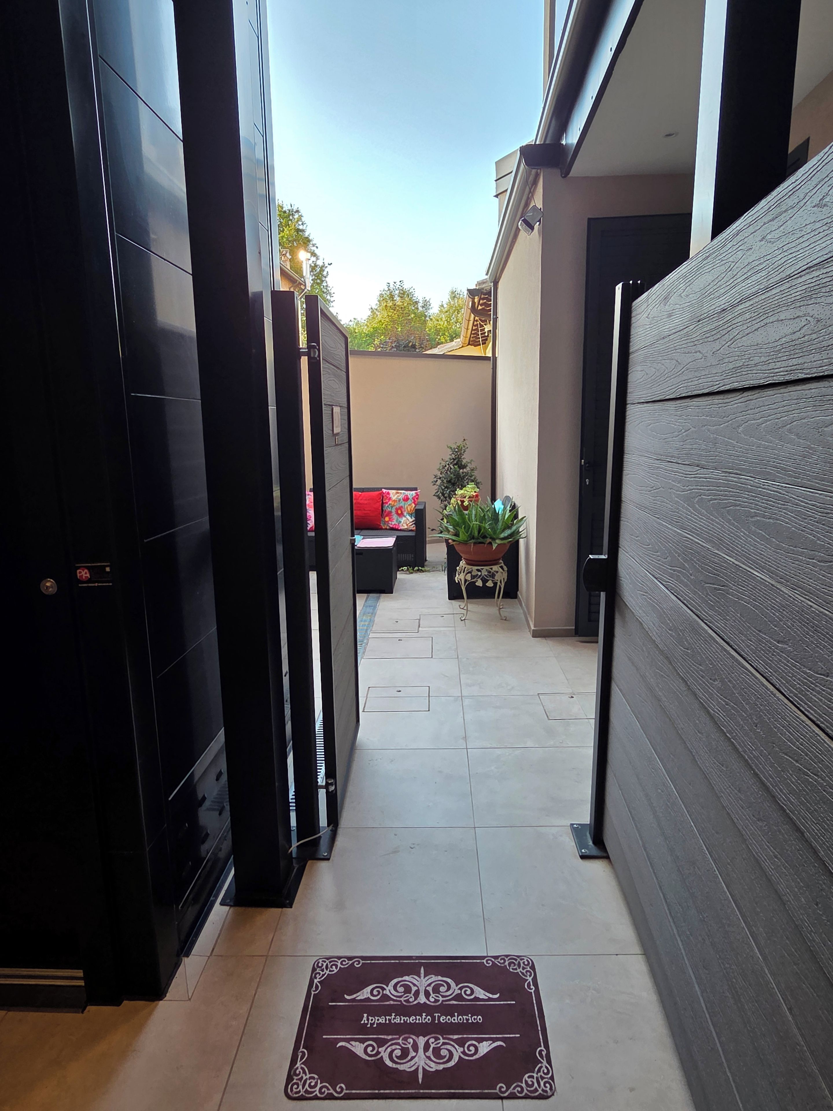 Corredor estreito com um tapete de entrada que diz 'Apartamento Teodorico', dando acesso a uma área de estar com um almofada vermelha e um vaso de planta.
