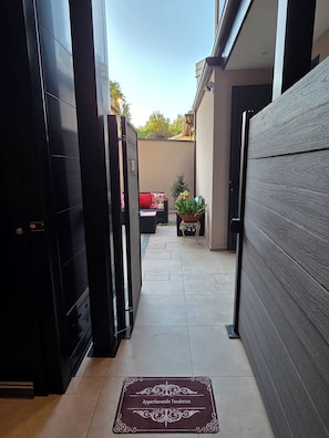 A narrow hallway with a doormat reading 'Apartamento Teodorico' leading to a seating area with a red cushion and a potted plant.