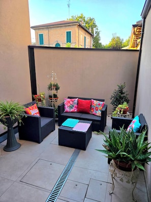 Un espacio al aire libre con mobiliario de mimbre, plantas en macetas y vista a edificios al fondo.