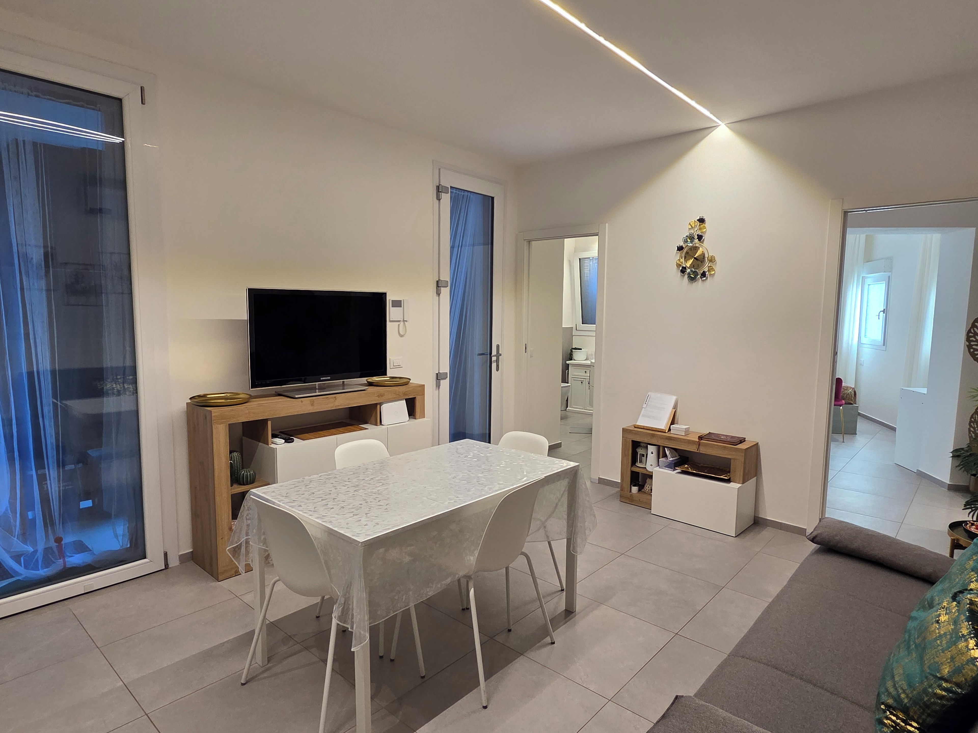 Ein modernes Wohnzimmer mit Essbereich, einem Flachbildfernseher auf einem Holzboden, einem weißen Tisch mit Stühlen und einem kleinen Beistelltisch mit einem Buch und einer Tasse.
