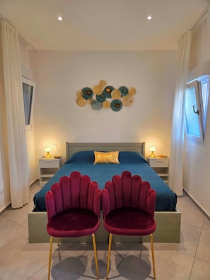 Un dormitorio con cama, dos sillas, mesitas de noche y un adorno en la pared.