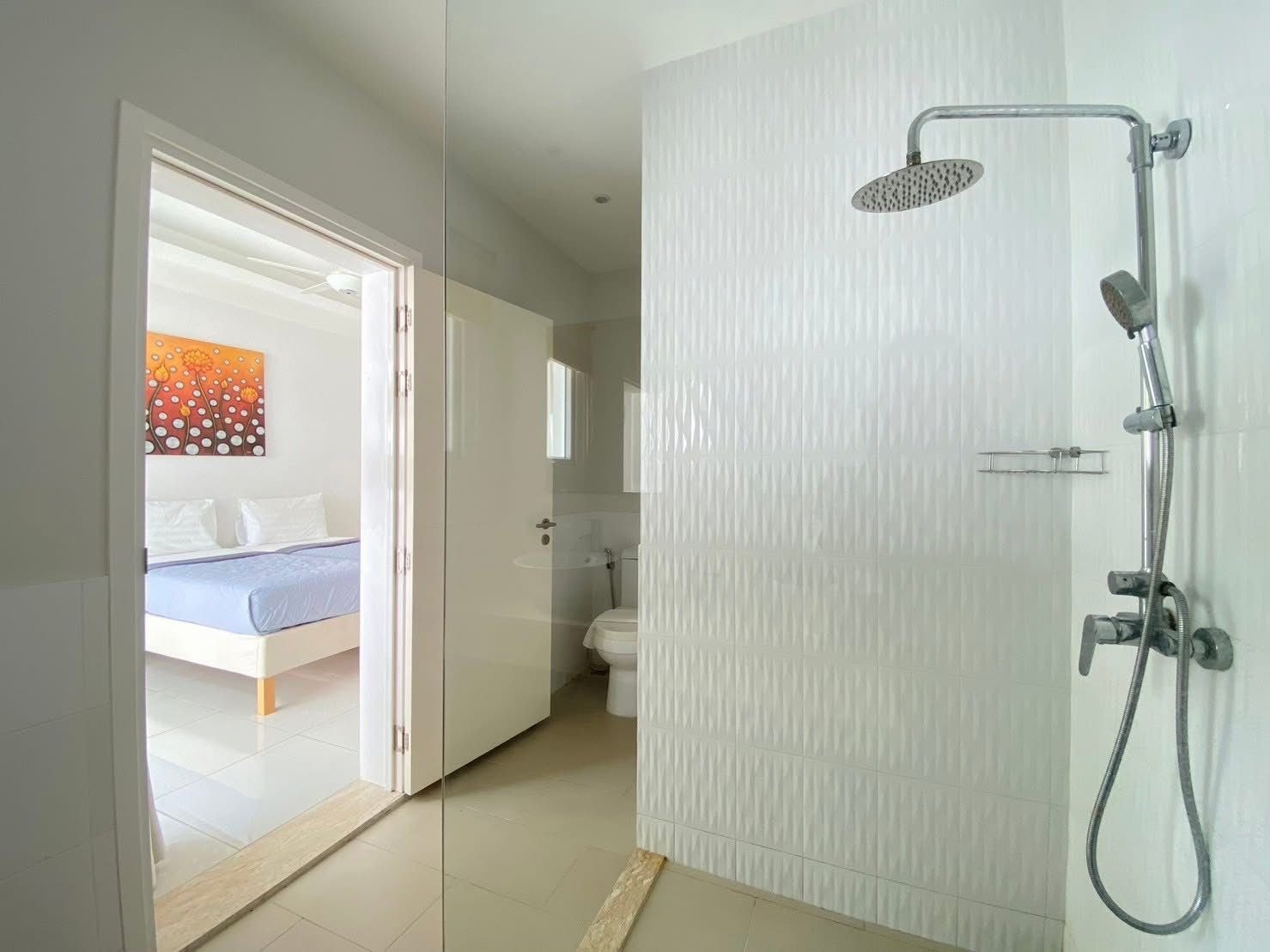 A modern bathroom with a shower, toilet, and a glass door leading to a bedroom with a bed and a painting.