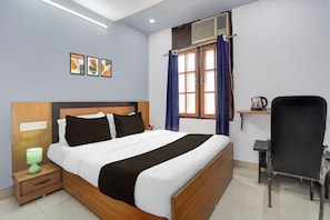 Une chambre d’hôtel comprenant un lit, une table de chevet, un bureau, une chaise, une fenêtre avec des rideaux et un mur orné de trois tableaux.