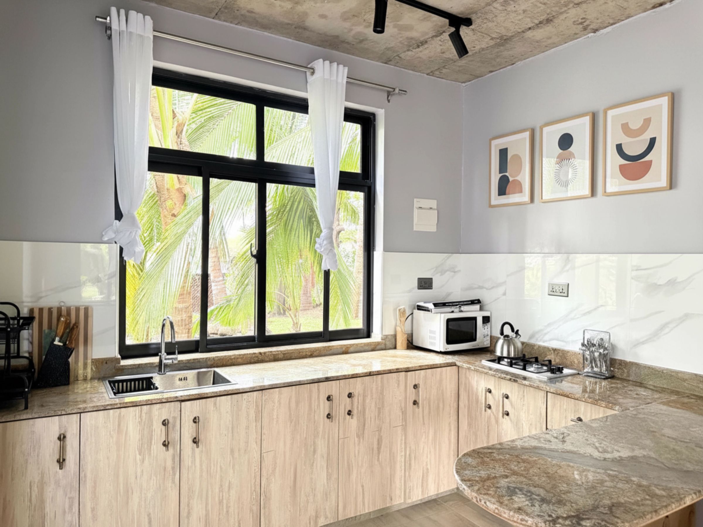A kitchen with a window, wooden cabinets, a sink, a microwave, and a kettle.