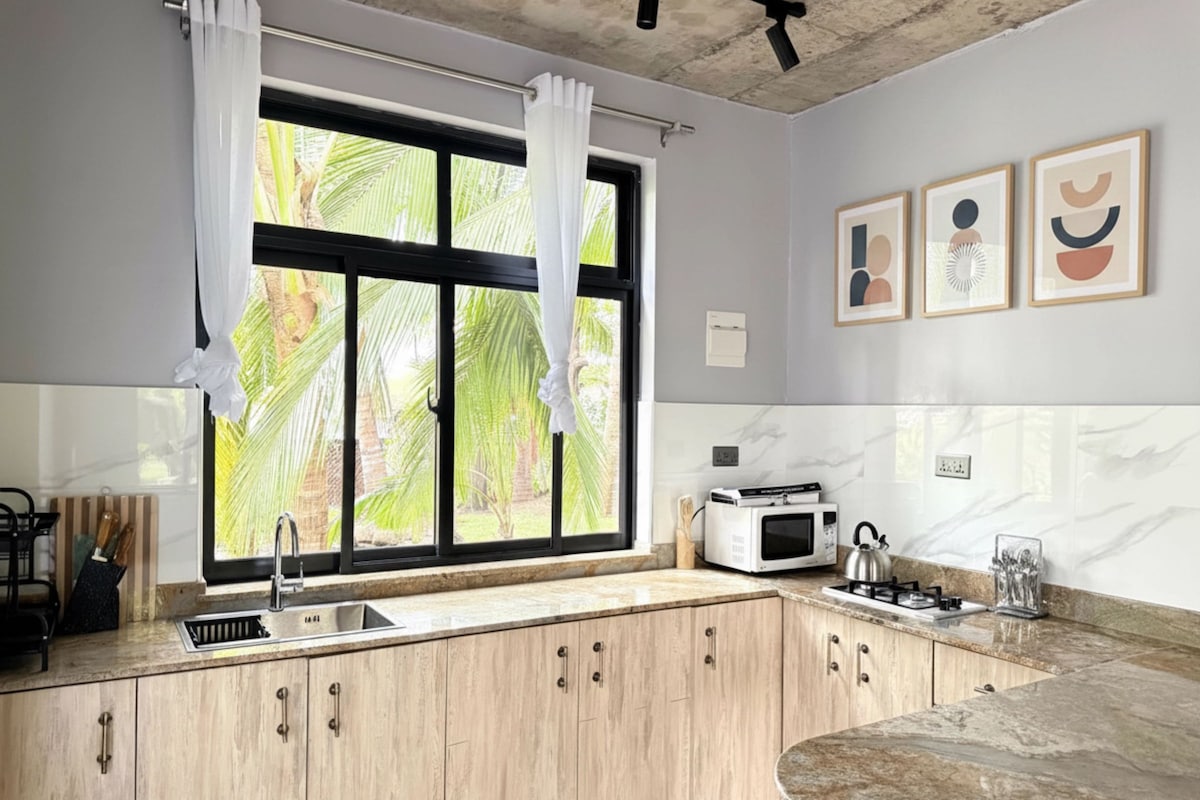 A kitchen with a window, wooden cabinets, a sink, a microwave, and a kettle.