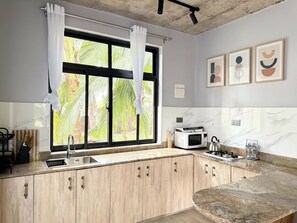 A kitchen with a window, wooden cabinets, a sink, a microwave, and a kettle.