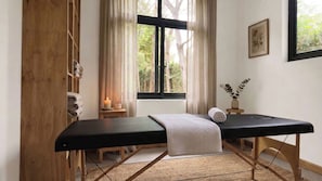 A room with a massage table, a window with a view of trees, a small bedside table with a candle, and a wall-mounted picture. - Nagira Tranquility (Taveta)