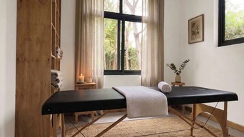Una habitación con una camilla de masaje, una ventana que da a árboles, una mesita de noche con una vela y un cuadro colgado en la pared.