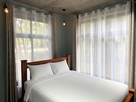 Quarto com cama de madeira, cortinas leves e luminária suspensa.