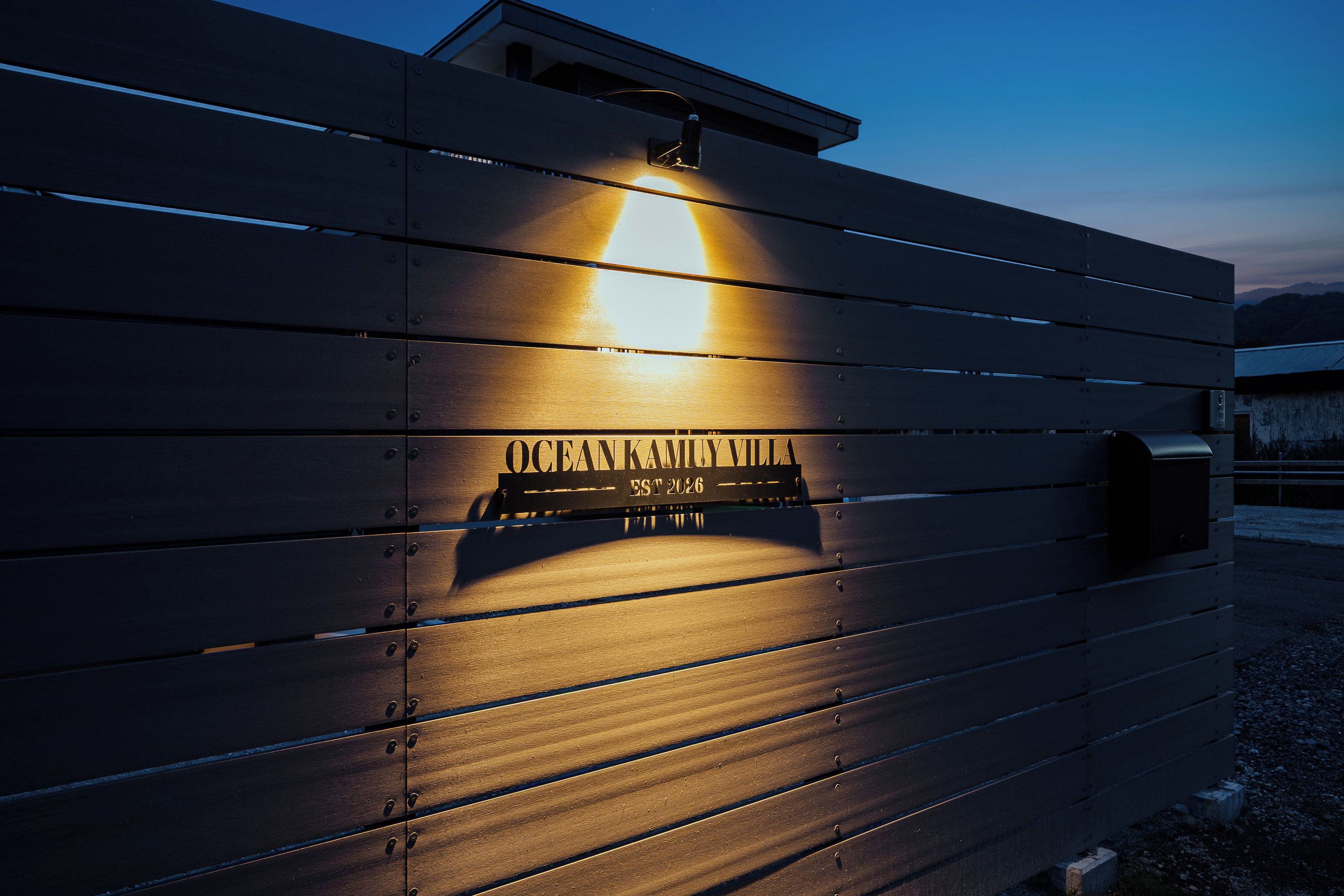 A wooden fence with a sign that reads 'OCEANAMYVILLA EST 2026' illuminated by a light.