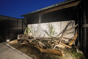 A modern building with a dark facade, a concrete pathway, and a landscaped area with logs and plants.