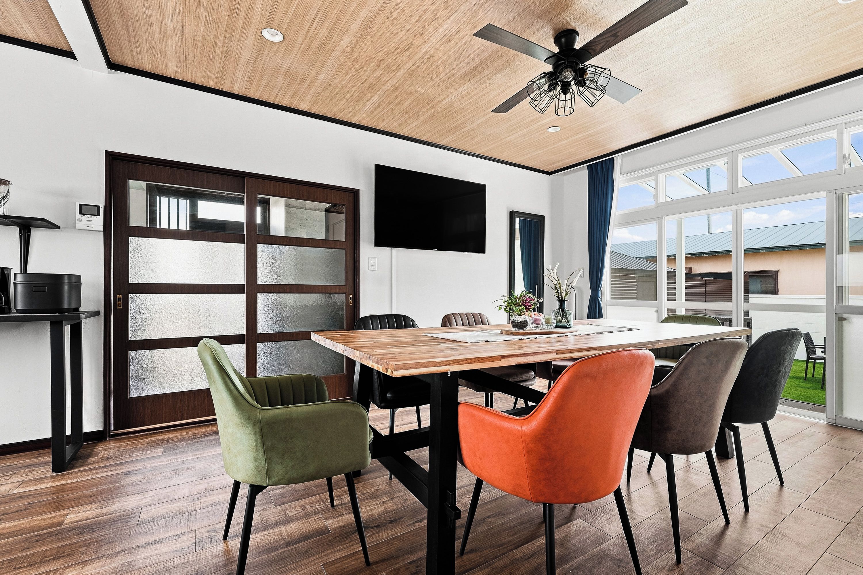 A modern dining area with a wooden table, colorful chairs, a ceiling fan, and a large window offering a view of the outdoors.