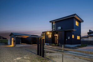 Une maison moderne à deux étages, avec une façade sombre, un climatiseur apparent et une entrée bien éclairée.