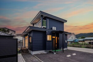 A modern two-story house with a balcony, a visible air conditioning unit, and a gated entrance.