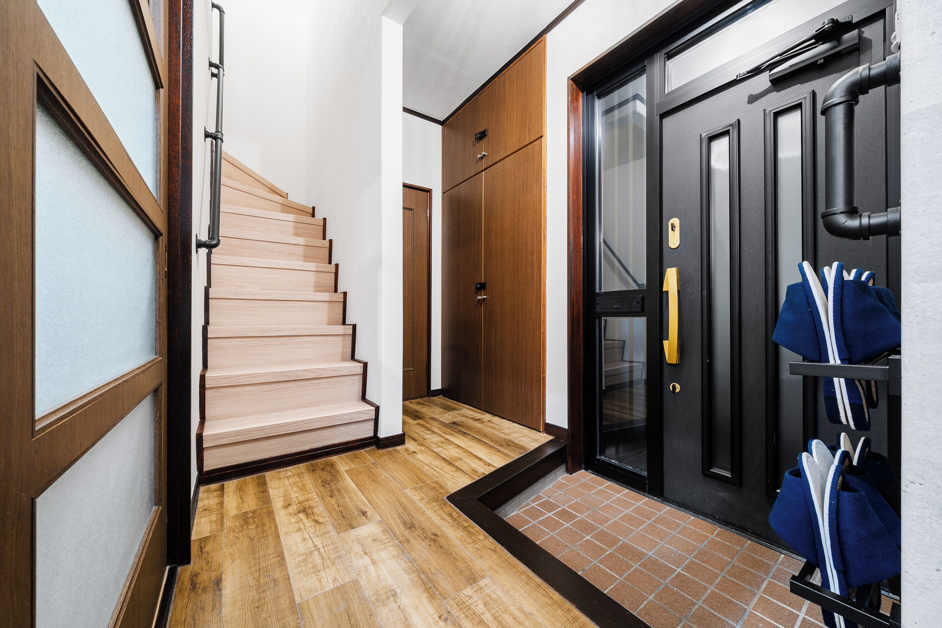 A hallway with a wooden staircase, a black door with a glass panel, and a shoe rack with shoes.