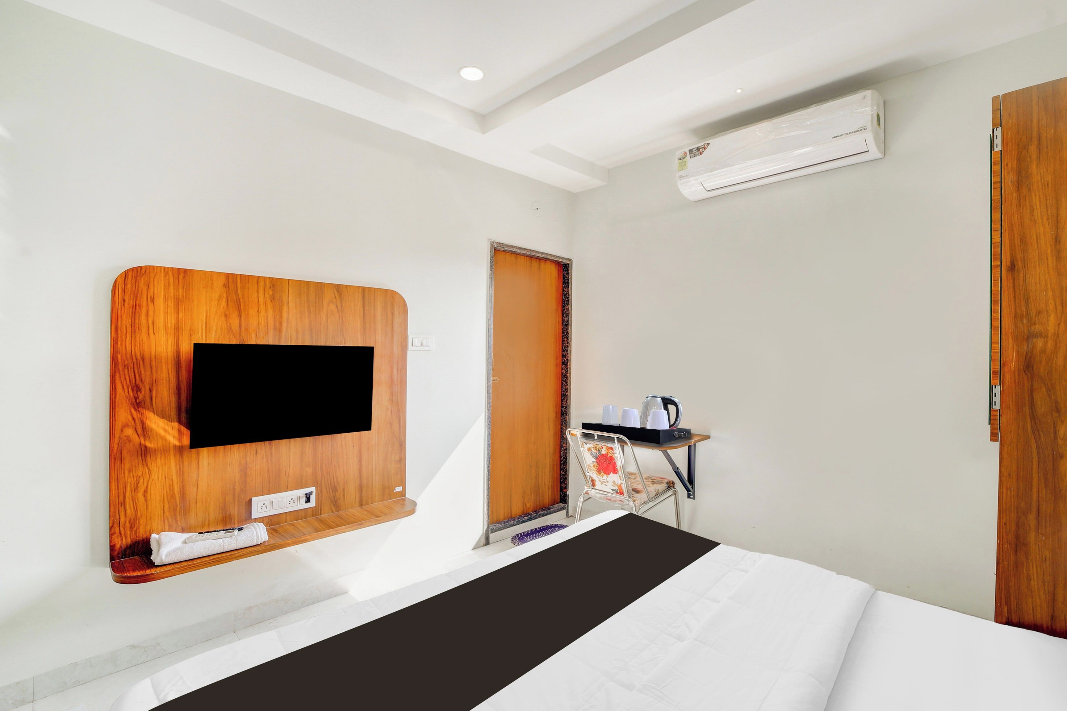 A hotel room with a bed, a TV mounted on the wall, an air conditioning unit, and a small table with a kettle and cups.