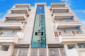 Un edificio de hotel de varios pisos con un ascensor de vidrio central, balcones en cada planta y un letrero que indica que es un hotel de servicios corporativos.