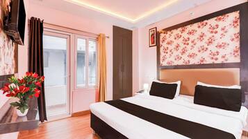 Une chambre d’hôtel comprenant un lit, une table de chevet, un téléphone, une lampe, une tête de lit décorée de motifs floraux, une fenêtre avec des rideaux et une plante.