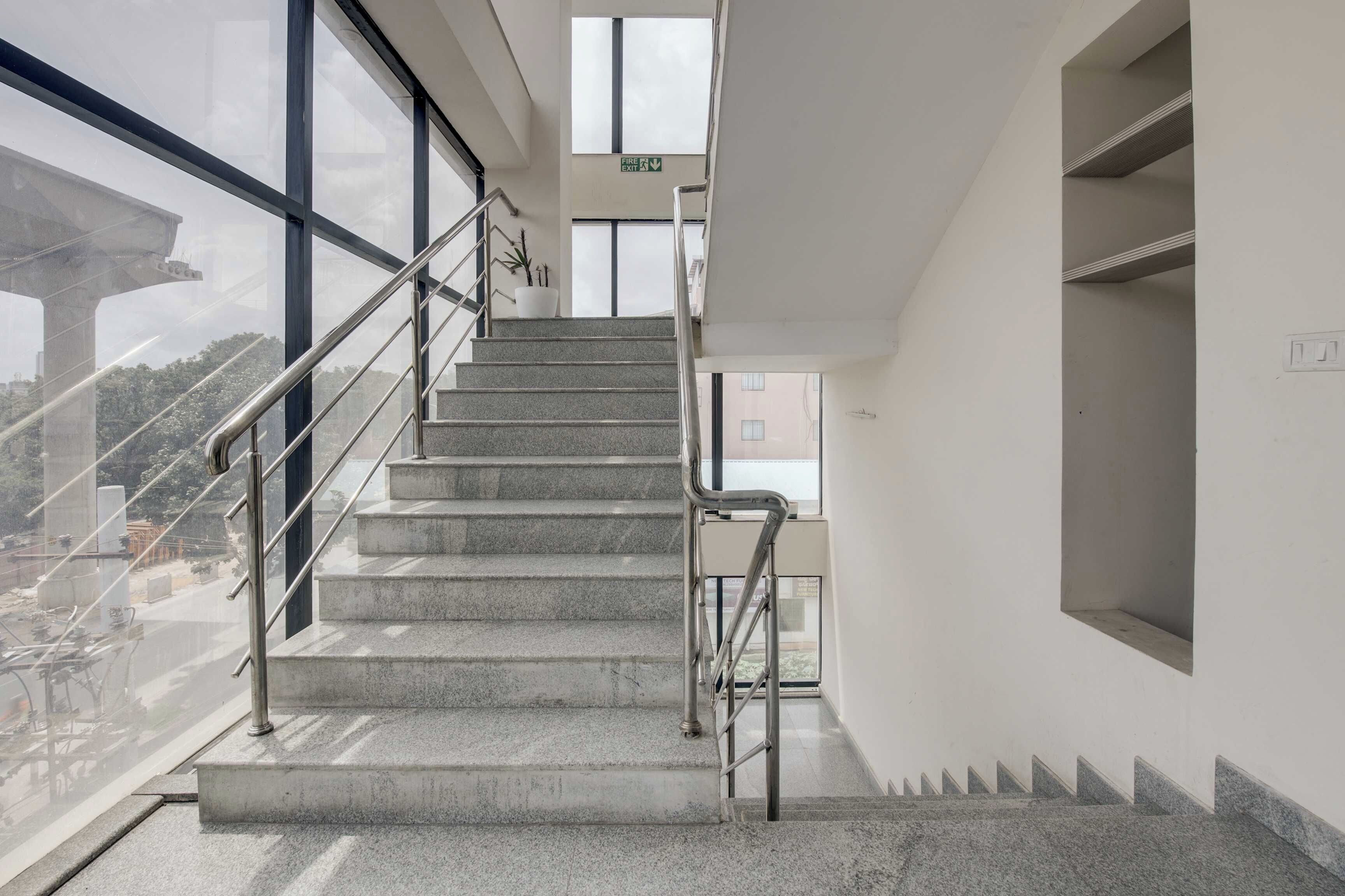 A staircase with metal railings leading to an upper floor, large windows on the left, and a fire exit sign above.