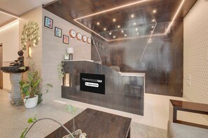 Una recepción con un cartel que indica servicio de la empresa, rodeada de un interior moderno con una pared de piedra, una estatua de Buda y una fuente de agua.