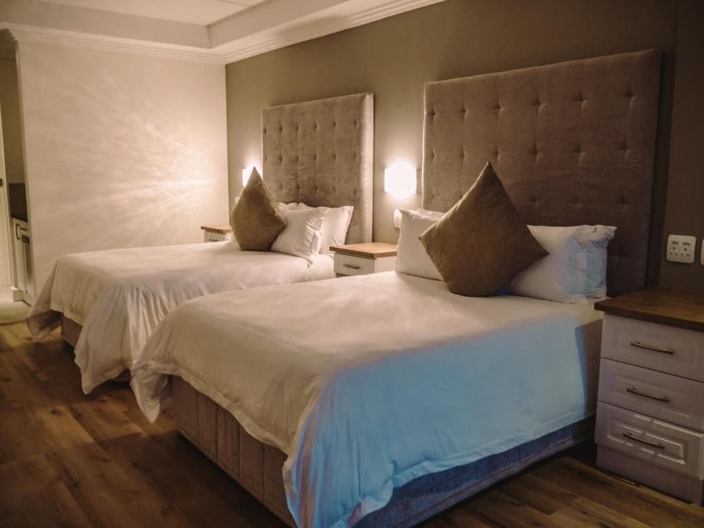 Habitación de hotel con dos camas, piso de madera y una mesita de noche con lámpara.