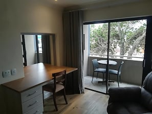 Una habitación con escritorio, silla y sofá. Hay una ventana con vista a árboles y un balcón.