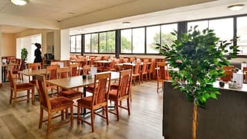 A spacious dining area with multiple wooden tables and chairs, a large potted plant, and a view through large windows.