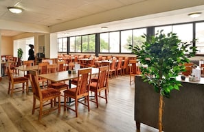 Een ruime eetruimte met meerdere houten tafels en stoelen, een grote potplant en uitzicht door grote ramen.